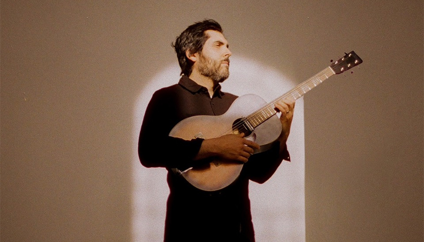 Ariel Posen With Guitar In Front Of A Beige Background