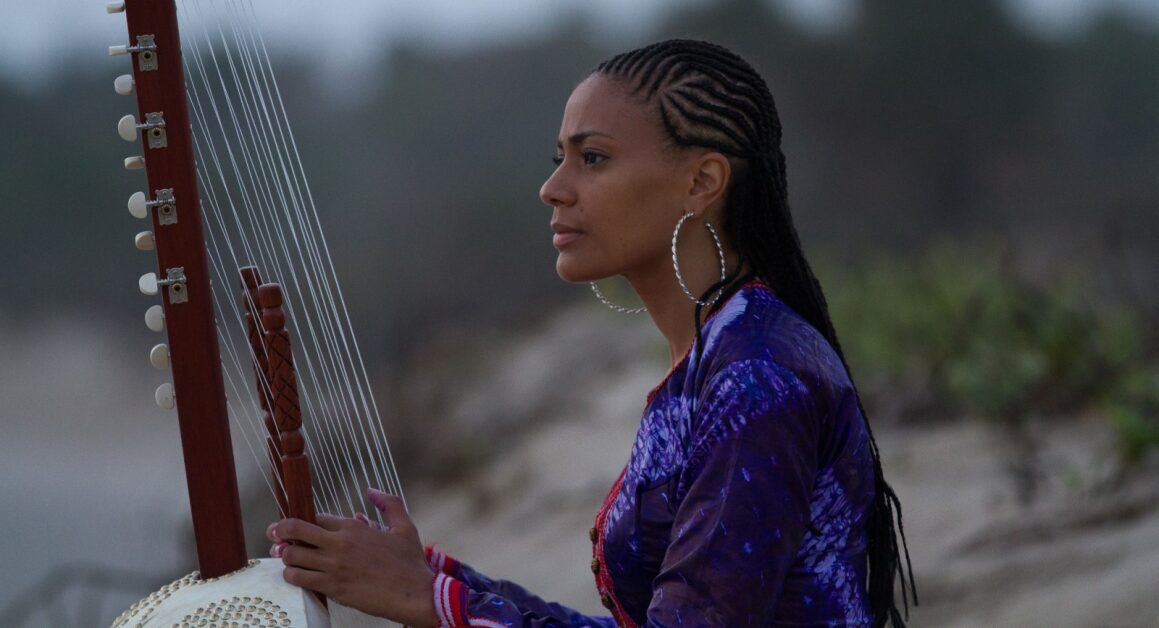 Sona Jobarteh Wearing A Blue Top With Her Hair In Braids Holding The Kora
