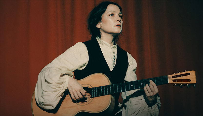 Natalia Lafourcade In A White Blouse And Black Vest Holding Her Guitar In Front Of A Red Curtain