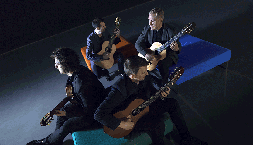 Dublin Guitar Quartet Wearing Suits And Holding Their Instruments While Sitting On A Futon