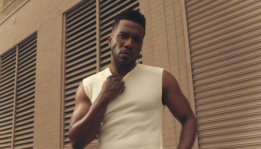 Leslie Odom Jr Pulling At The Collar Of A White Tank Top In Front Of A Brown Building