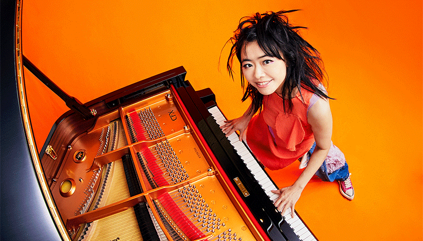 Hiromi In An Orange Shirt And Jeans Playing A Grand Piano