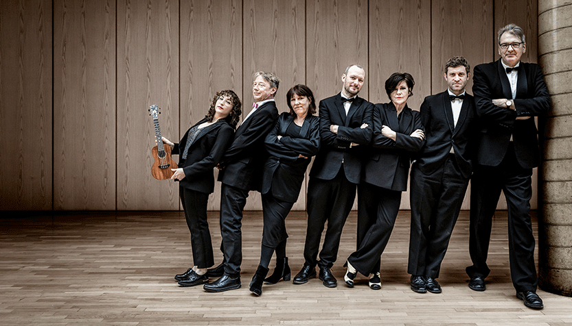 Ukulele Orchestra Of Great Britain Wearing Tuxedos And Leaning Against Each Other