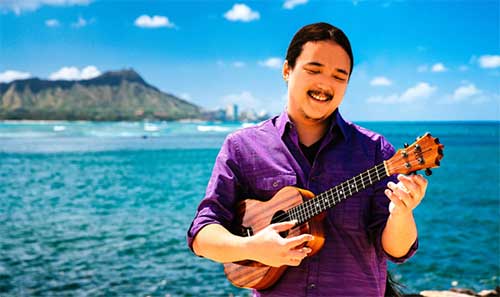 Neal Chin Wearing A Purple Shirt Holding His Ukulele In Front Of The Hawaiian Coast